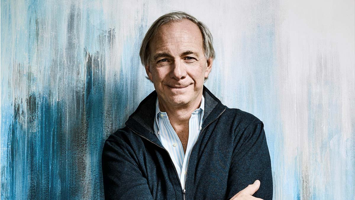 Älterer Mann mit grauen Haaren und blauem Pullover vor künstlerisch gestrichenem Hintergrund, verschränkt die Arme und lächelt